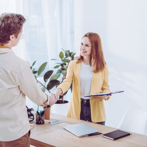 happy-recruiter-holding-clipboard-and-shaking-hands-with-employee-in-modern-office.jpg